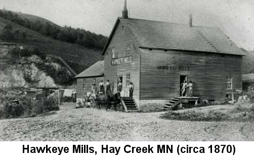 Hawkeye Mills, Hay Creek, MN (circa 1870); black and white photo of a frame mill building with several people standing on its two porches, and wooded hills in the background